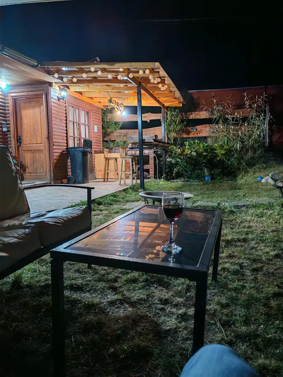 La cabaña de madera de noche, con el quincho iluminado y una copa de vino en la mesa